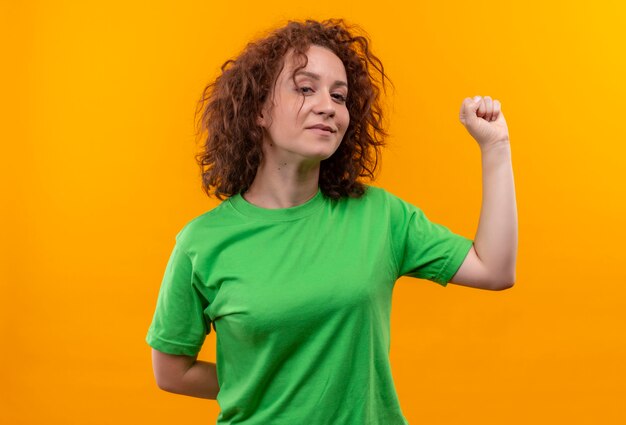 Giovane donna con i capelli ricci corti in maglietta verde che alza il pugno che sembra fiducioso in piedi sopra la parete arancione