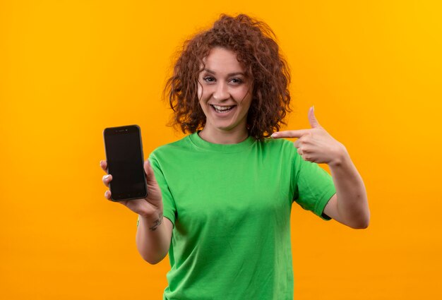 Giovane donna con i capelli ricci corti in maglietta verde che mostra lo smartphone che punta con il dito sorridendo allegramente in piedi sopra la parete arancione