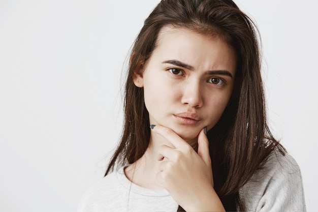 Giovane donna con i capelli scuri e gli occhi scuri che aggrotta le sopracciglia tenendo il dito indice sul mento con uno sguardo dubbioso e sospettoso, scettico su qualcosa. Emozioni umane e concetto di espressione del viso