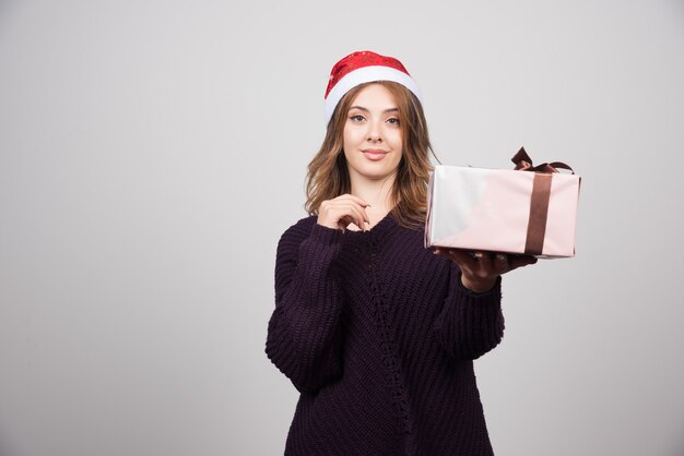 Giovane donna con il cappello di Babbo Natale che mostra un regalo festivo con fiocco.