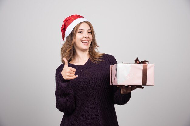 Giovane donna con il cappello di Babbo Natale con un regalo che mostra un pollice in su.