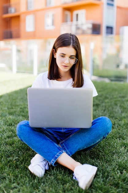 Giovane donna con il computer portatile che si siede sull'erba verde e che guarda ad un'esposizione all'aperto