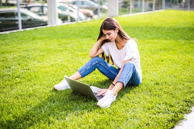 Giovane donna con il computer portatile che si siede sull'erba verde e che osserva ad un'esposizione