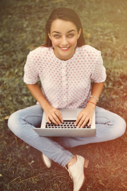 Giovane donna con il computer portatile che si siede sull'erba verde