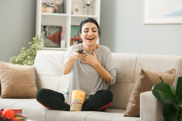 Giovane donna con il secchio di popcorn che tiene il telecomando della tv alla telecamera seduta sul divano dietro il tavolino da caffè in soggiorno