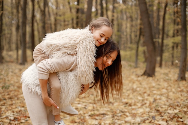 Giovane donna con la bambina in piedi nella foresta d'autunno. Donna castana che tiene sua figlia sulle spalle. Ragazza che indossa un maglione beige e madre che indossa abiti bianchi.