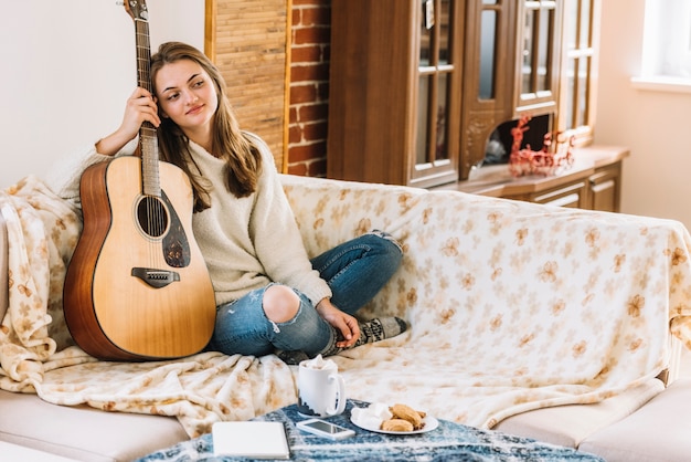 Giovane donna con la chitarra vicino tavolo con smartphone, blocco note, tazza di bevanda e biscotti