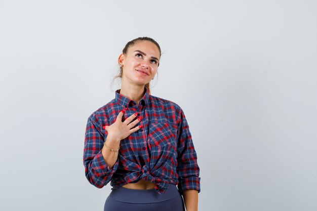 Giovane donna con la mano sul petto in camicia a scacchi, pantaloni e dall'aspetto carino. vista frontale.