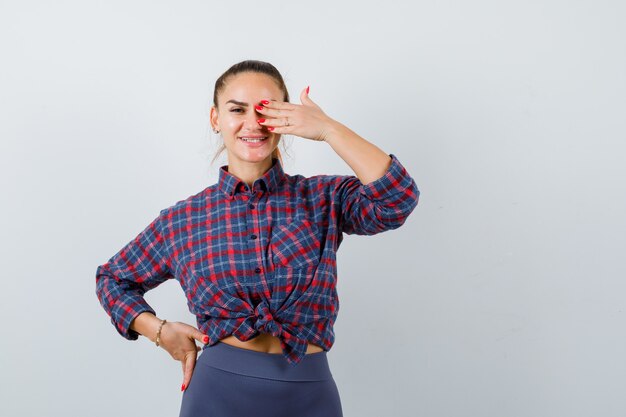 Giovane donna con la mano sull'occhio mentre tiene la mano sull'anca in camicia a scacchi, pantaloni e sembra felice. vista frontale.