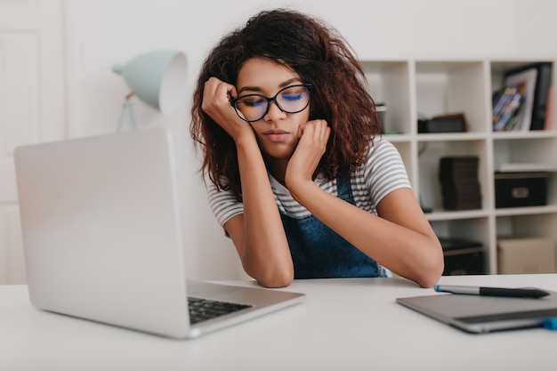 Giovane donna con la pelle marrone chiaro indossa occhiali alla moda che dorme davanti al computer portatile in ufficio