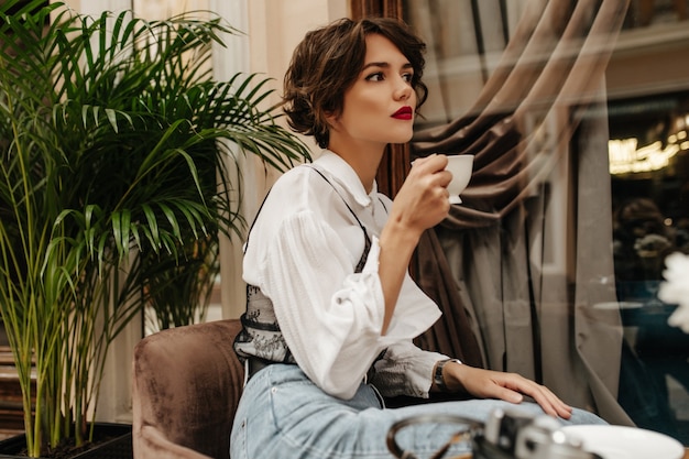 Giovane donna con labbra luminose e capelli ricci pone nel ristorante. Donna alla moda in jeans e camicia bianca tiene la tazza di caffè nella caffetteria.