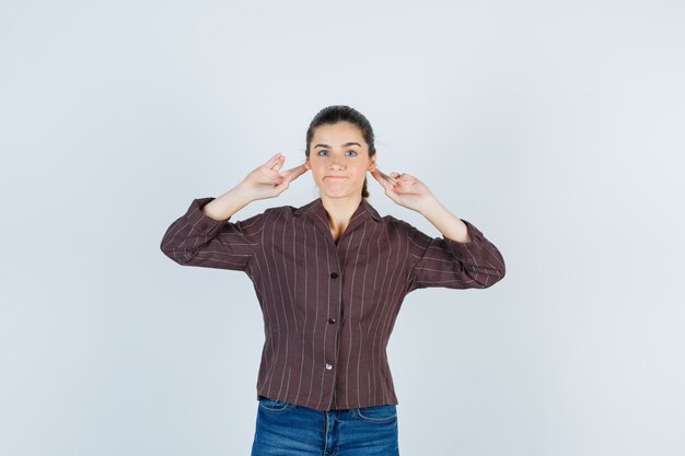 Giovane donna con le mani dietro le orecchie in camicia a righe, jeans e dall'aspetto carino, vista frontale.