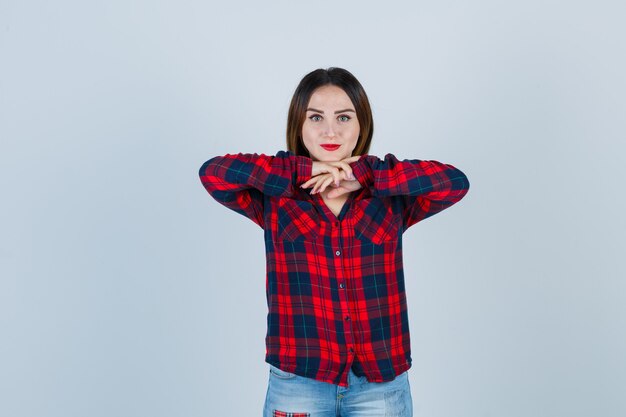 Giovane donna con le mani sotto il mento, mento appoggiato sulle mani in camicia a quadri, jeans e aspetto attraente. vista frontale.