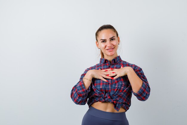 Giovane donna con le mani sul petto in camicia a scacchi, pantaloni e guardando allegro, vista frontale.