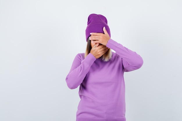 Giovane donna con le mani sul viso in maglione viola, berretto e guardando addolorato. vista frontale.
