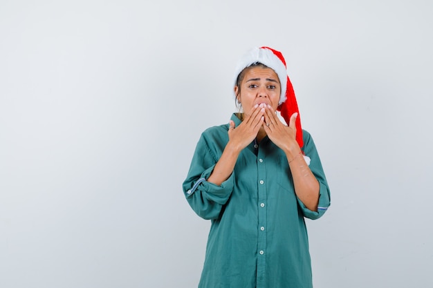 Giovane donna con le mani sulla bocca in cappello di natale, camicia e sembra spaventata. vista frontale.