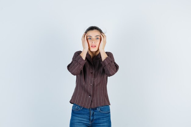 Giovane donna con le mani sulle tempie in camicia a righe, jeans e guardando sorpreso, vista frontale.