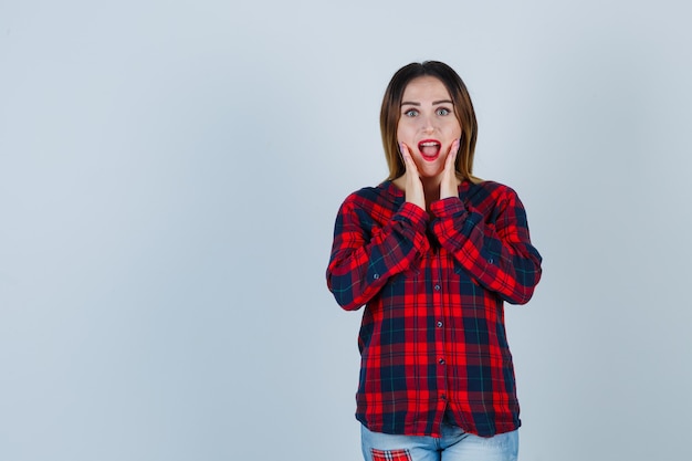 Giovane donna con le mani vicino alla bocca aperta in camicia a quadri e guardando stupito, vista frontale.