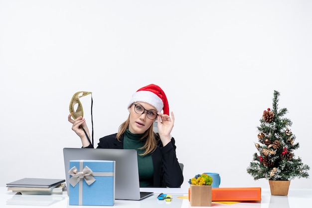 Giovane donna con occhiali cappello di Babbo Natale e tenendo la maschera seduto a un tavolo con un albero di Natale e un regalo