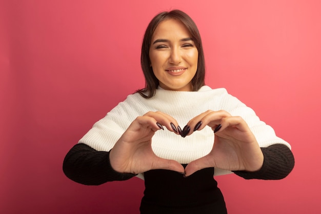 Giovane donna con sciarpa bianca che fa il gesto del cuore con le dita che sorridono allegramente