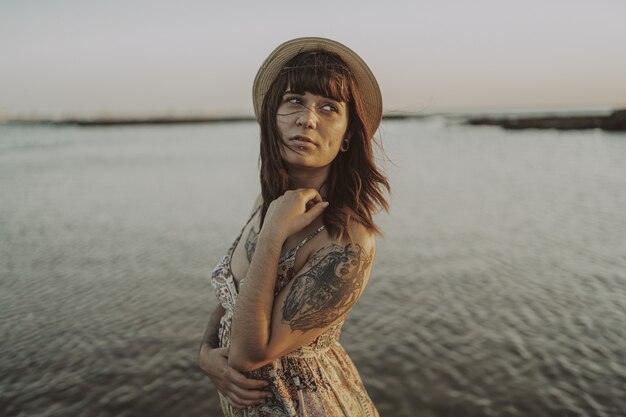 Giovane donna con tatuaggi che indossa un vestito e un cappello di paglia in spiaggia