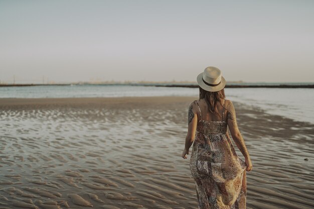 Giovane donna con tatuaggi che indossa un vestito e un cappello di paglia sull'oceano sfocato