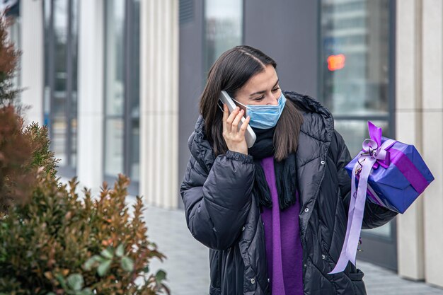 Giovane donna con una maschera protettiva con un regalo in mano che parla al telefono