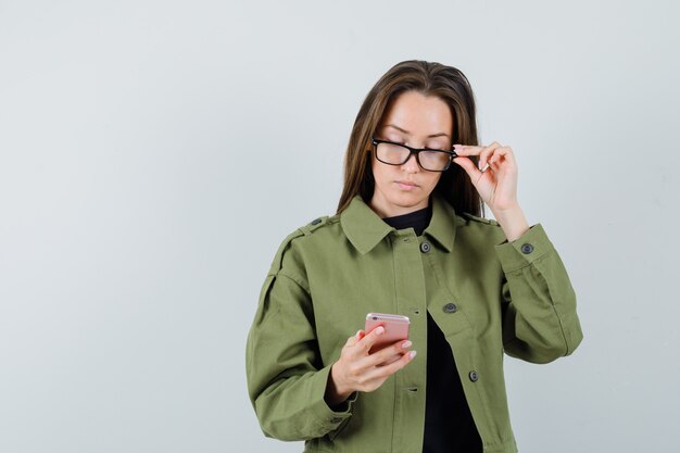 Giovane donna concentrandosi sul suo telefono cellulare in giacca verde, occhiali, vista frontale.