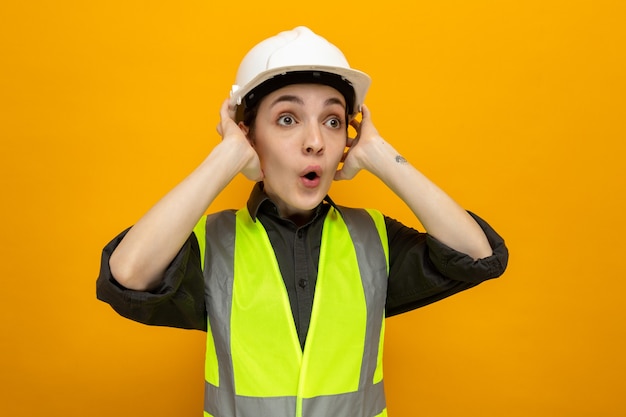 Giovane donna costruttore in giubbotto da costruzione e casco di sicurezza che guarda da parte stupita e sorpresa tenendosi per mano sulla testa in piedi sul muro arancione