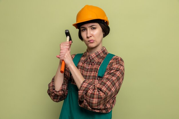 Giovane donna costruttore in uniforme da costruzione e casco di sicurezza che tiene martello con faccia seria in piedi sul verde