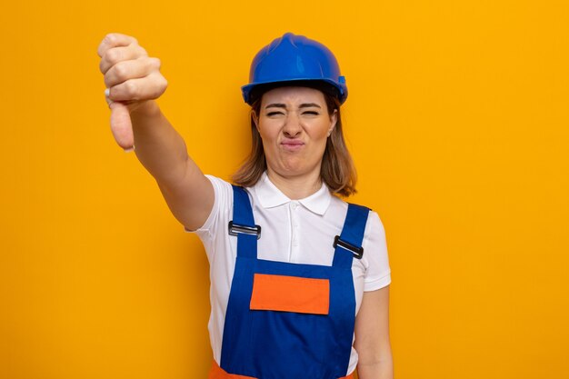 Giovane donna costruttore in uniforme da costruzione e casco di sicurezza dispiaciuto che mostra i pollici verso il basso in piedi sul muro arancione