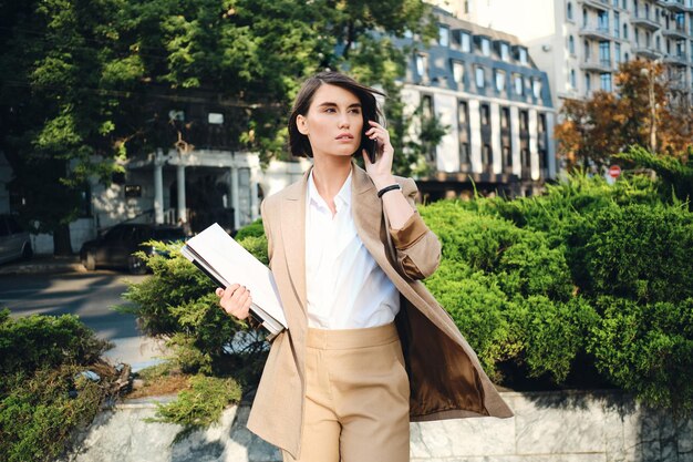 Giovane donna d'affari attraente in abito beige con il computer portatile che parla pensierosamente sul cellulare sulla strada della città