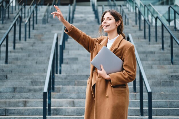 Giovane donna d'affari casual sorridente in cappotto con il computer portatile agitando con gioia la mano ciao gesto all'aperto