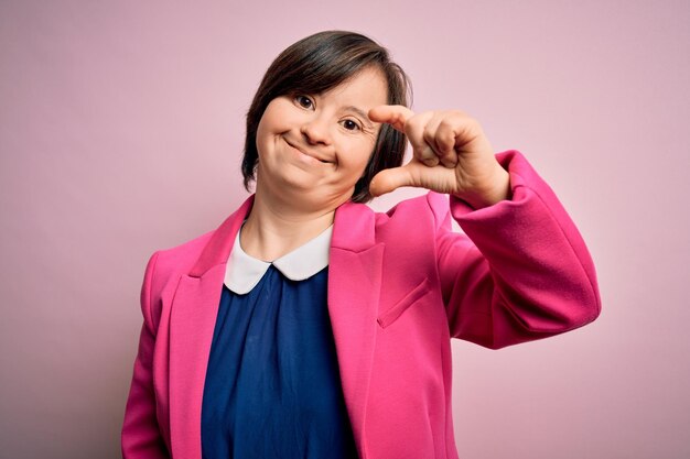 Giovane donna d'affari con sindrome di Down su sfondo rosa sorridente e gesticolando fiducioso con la mano facendo un segno di piccole dimensioni con le dita che guardano e la fotocamera Concetto di misura