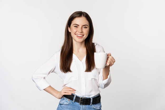 Giovane donna d'affari modera, signora dell'ufficio che tiene tazza con caffè, tè e sorridente, in piedi su sfondo bianco