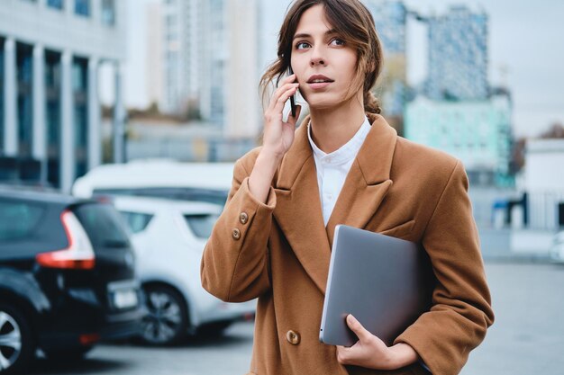 Giovane donna d'affari pensierosa in cappotto con il laptop che parla al cellulare mentre distoglie lo sguardo sulla strada della città