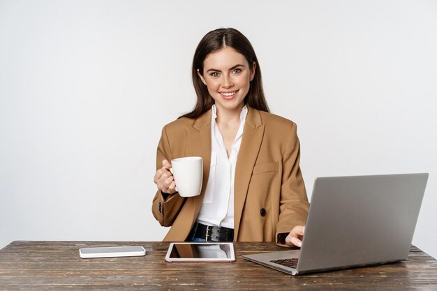 Giovane donna d'affari professionale nel suo ufficio, seduta vicino al laptop e sorridente, bevendo caffè, posando su sfondo bianco