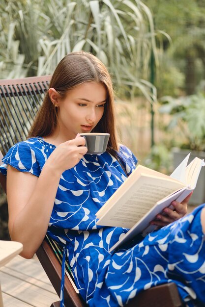 Giovane donna dai capelli castani attraente in abito blu che beve caffè e legge premurosamente un libro sulla sedia a sdraio di legno nel parco cittadino