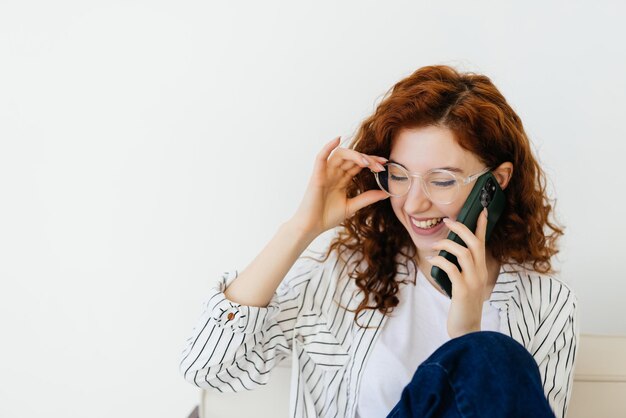 Giovane donna dai capelli rossi seduta a casa divano divano a bere caffè parlando al telefono cellulare rilassata nel concetto di comunicazione e stile di vita