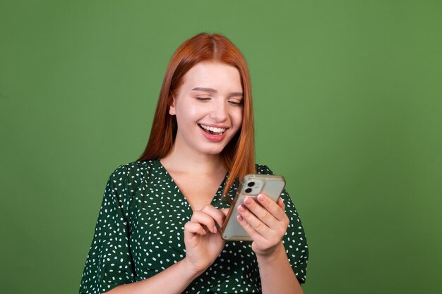 Giovane donna dai capelli rossi sulla parete verde con il telefono cellulare felice positivo eccitato