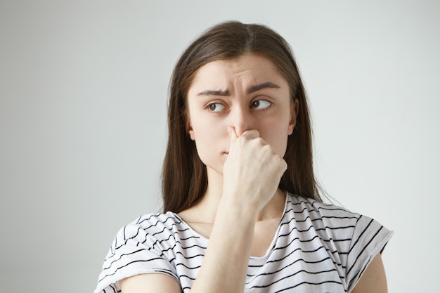 Giovane donna dai capelli scura e frustrata isolata orizzontale con sguardo disgustato, pizzicandosi il naso e trattenendo il respiro a causa di odore, odore o puzza sgradevole