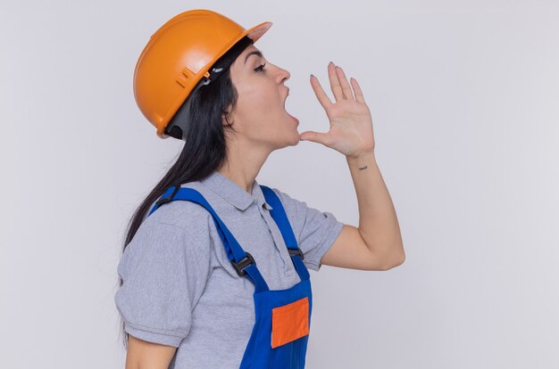 Giovane donna del costruttore in uniforme da costruzione e casco di sicurezza che grida o chiama qualcuno che hlding consegna la bocca in piedi sopra il muro bianco
