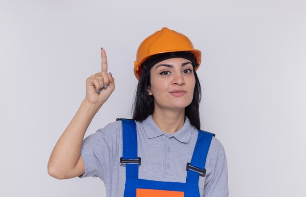 Giovane donna del costruttore in uniforme da costruzione e casco di sicurezza che guarda davanti con il sorriso sul viso intelligente che mostra il dito indice in piedi sul muro bianco