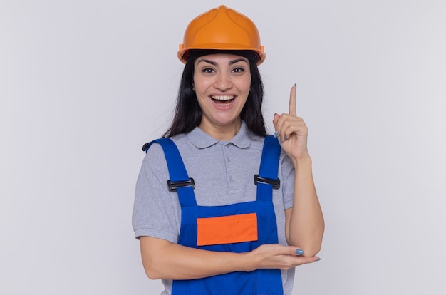 Giovane donna del costruttore in uniforme da costruzione e casco di sicurezza che guarda davanti con il sorriso sul viso intelligente che mostra il dito indice in piedi sul muro bianco