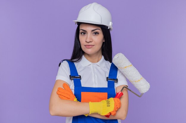 Giovane donna del costruttore in uniforme da costruzione e casco di sicurezza che indossa guanti di gomma che tengono il rullo di vernice guardando davanti con espressione seria e sicura in piedi sopra la parete viola