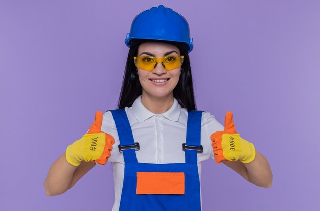 Giovane donna del costruttore in uniforme da costruzione e casco di sicurezza che indossa guanti di gomma