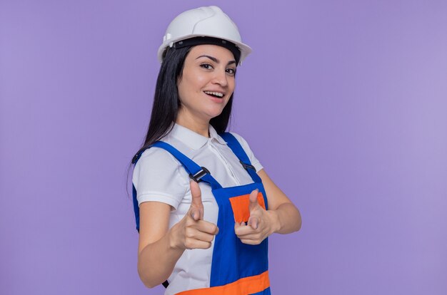 Giovane donna del costruttore in uniforme da costruzione e casco di sicurezza che sorride allegramente indicando con le dita indice davanti in piedi sopra la parete viola