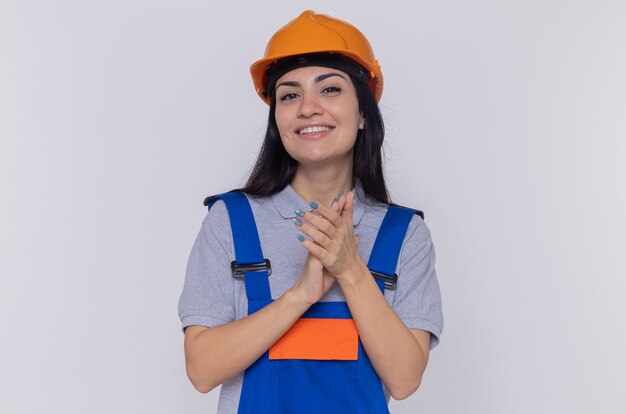 Giovane donna del costruttore in uniforme da costruzione e casco di sicurezza che tengono le mani insieme guardando la parte anteriore sorridente fiducioso in piedi sopra il muro bianco