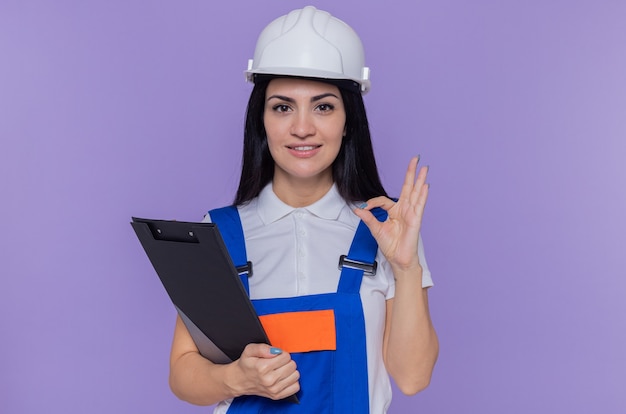 Giovane donna del costruttore in uniforme da costruzione e casco di sicurezza che tiene appunti e matita guardando davanti sorridente felice e positivo fiducioso facendo segno giusto in piedi sopra il muro viola