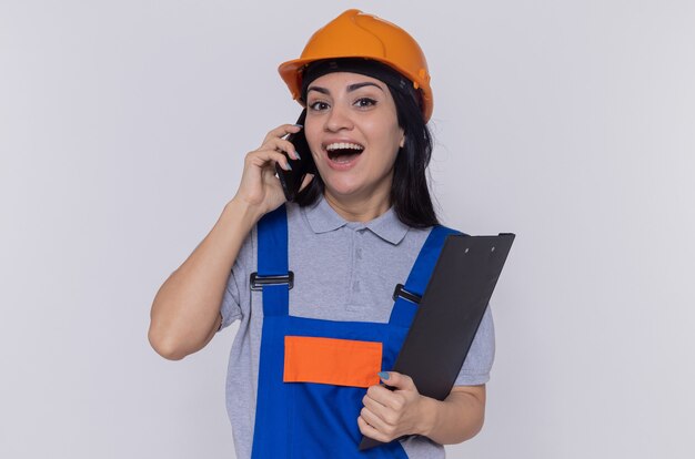 Giovane donna del costruttore in uniforme da costruzione e casco di sicurezza che tiene appunti sorridendo allegramente mentre parla al telefono cellulare in piedi sopra il muro bianco